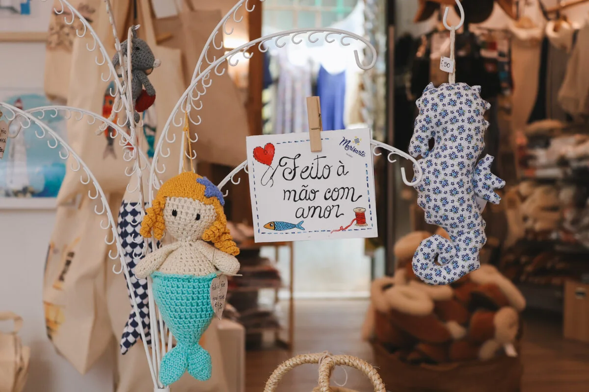 Uma sereia de croché com cabelo amarelo, um cavalo-marinho de tecido azul e branco e a etiqueta escrita à mão «Feito à mão com amor»